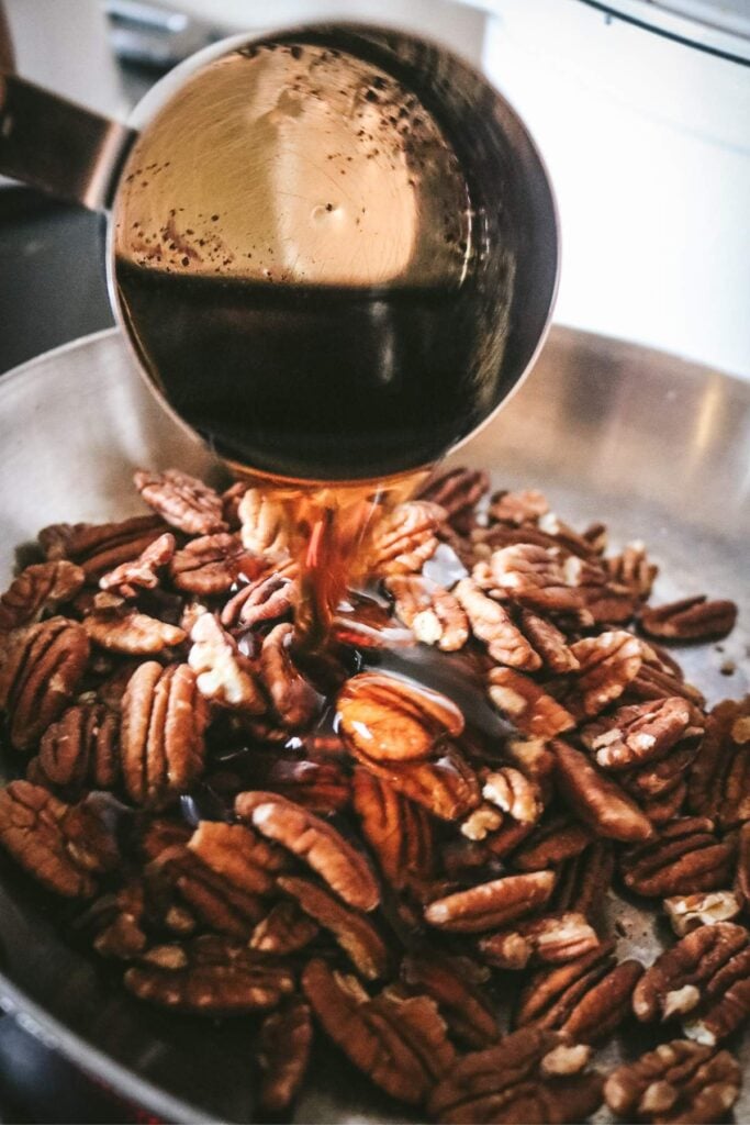pouring maple syrup over pecans in a skillet.