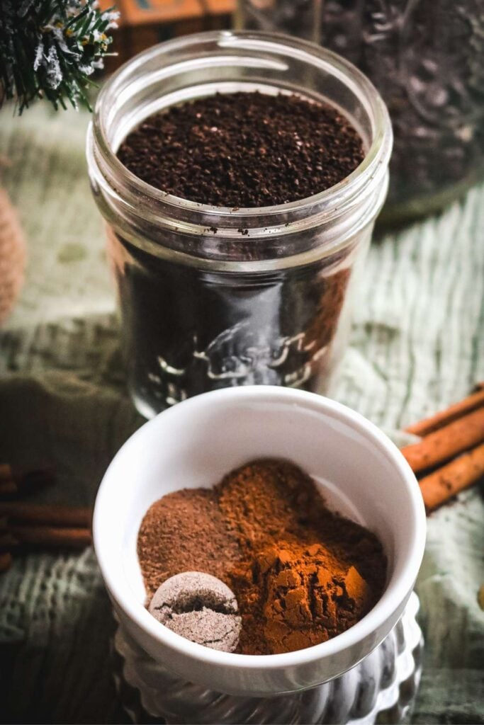 glass mason jar with freshly ground coffee and a white dish with different spices like cinnamon, cardamom, and more.
