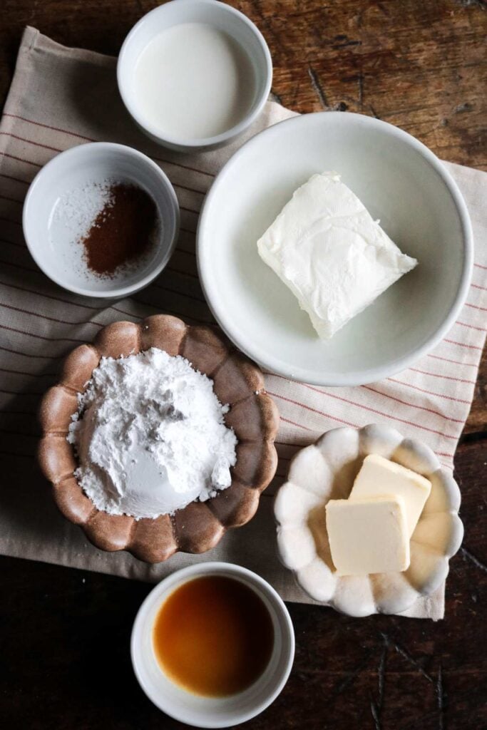 overhead view of different bowls showing the ingredients to make cinnamon cream cheese frosting like powdered sugar, vanilla extract, and butter.