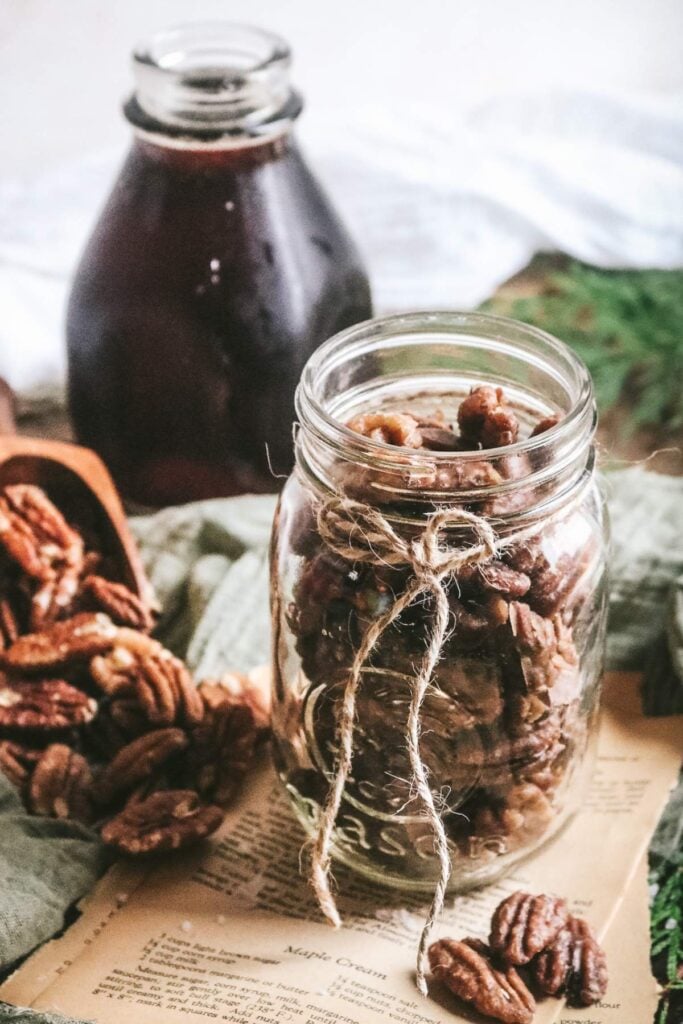 a mason jar with candied maple pecans next to pecans and a bottle of maple syrup.