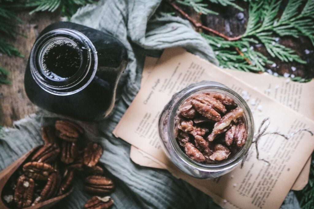 overhead view of a homemade maple pecans by cedar boughs, pecans, and a bottle of maple syrup.