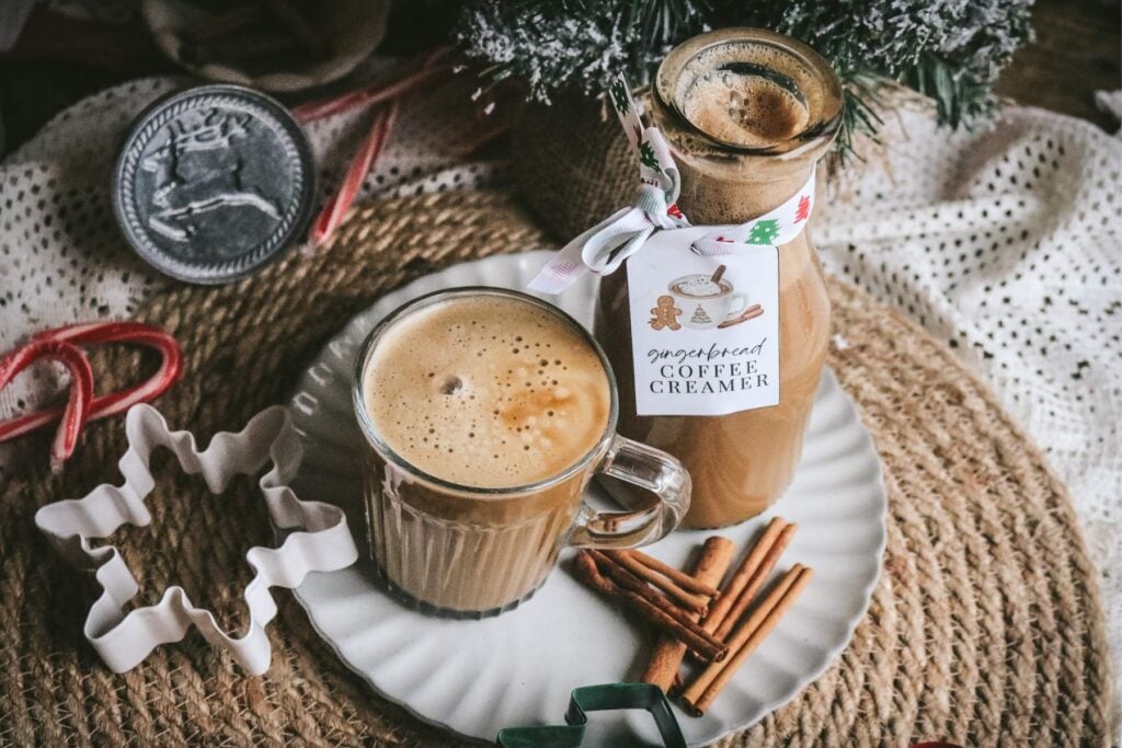 overhead view of a wooden table with homemade gingerbread coffee creamer in a glass bottle next to a mug of coffee and various holiday accessories, like cookie cutters.