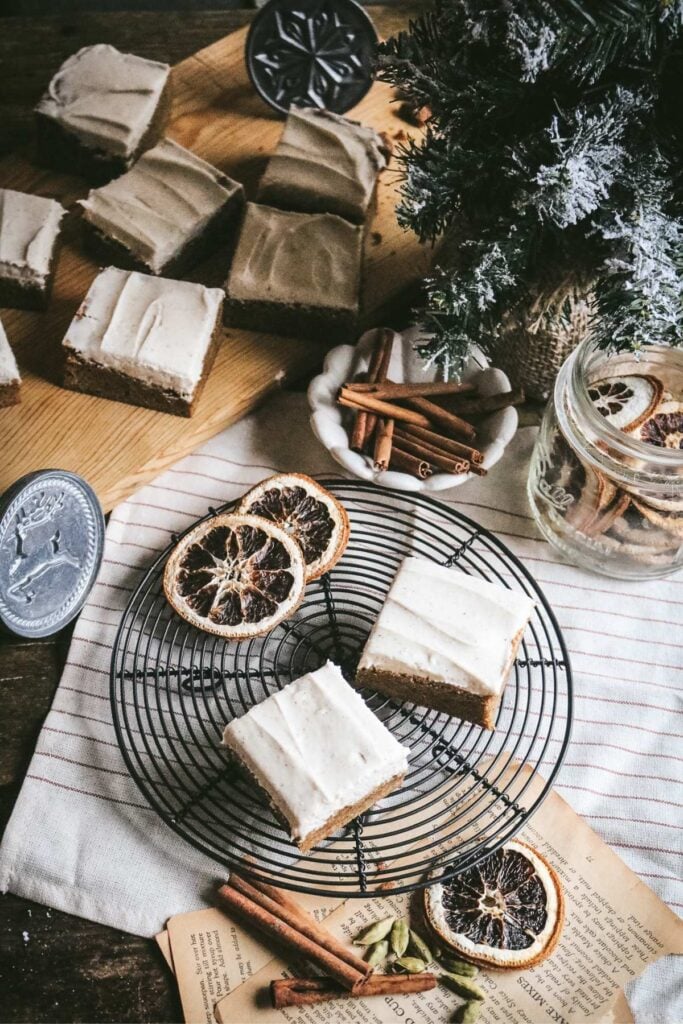 wooden table with an antique wire rack holding dried orange slices and frosted gingerbread blondies with cinnamon sticks, a christmas tree, and more blondies.