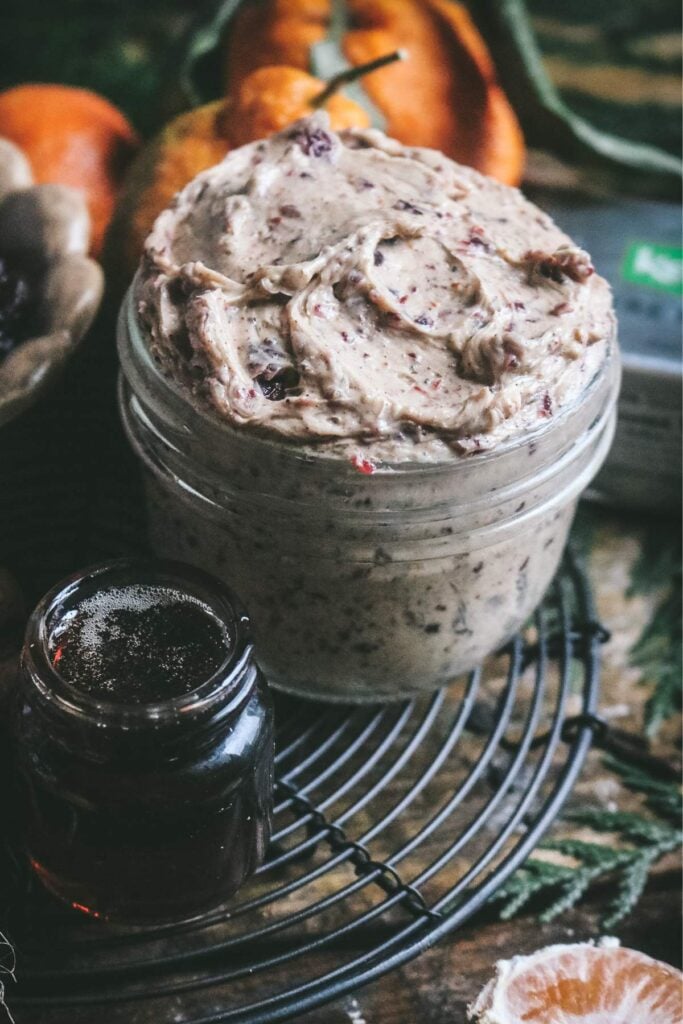 jar of thick, creamy cinnamon cranberry compound butter with oranges in the back and maple syrup in the front.