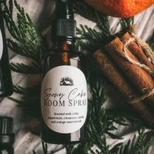 image of an amber glass spray bottle with a white label that says snowy cabin room spray with cedar, cinnamon sticks, and orange in the background.