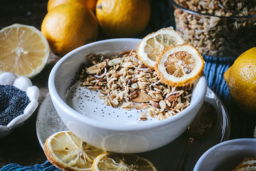 white bowl with yogurt, homemade lemon poppyseed granola, and dried lemon slices surrounded by lemons, a bowl of poppyseeds, and extra granola.