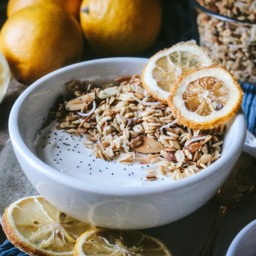 white bowl with yogurt, homemade lemon poppyseed granola, and dried lemon slices surrounded by lemons, a bowl of poppyseeds, and extra granola.