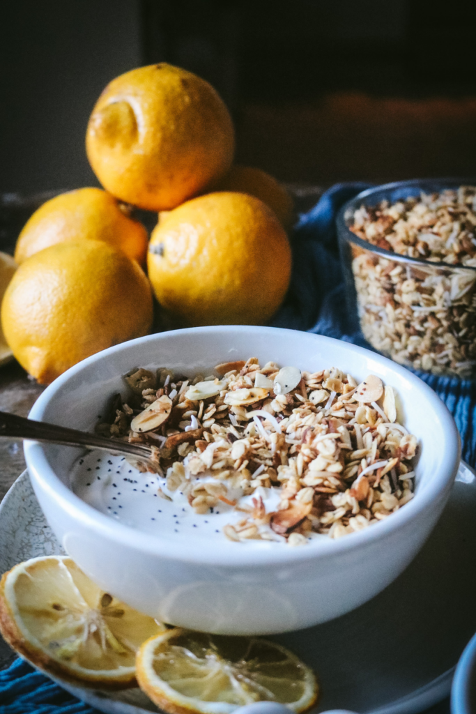 Greek yogurt with a serving of lemon poppyseed granola with coconut shreds and almond next to fresh lemons and dried lemon slices.