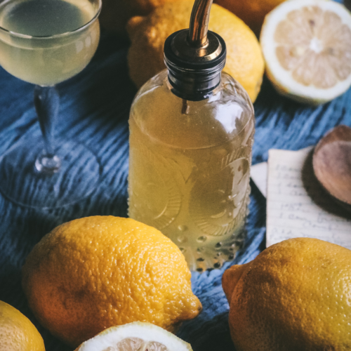 glass bottle of homemade lemon simple syrup surrounded by fresh lemons on a blue cloth.