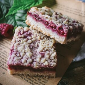 close up view of homemade raspberry crumble bars on vintage book pages in front of fresh raspberries and raspberry leaves.