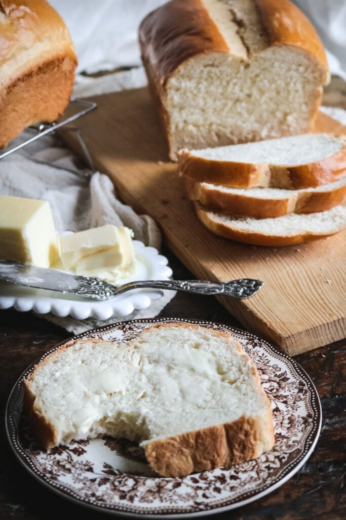 close up view of a slice of homemade sandwich bread slathered with butter on an antique brown transferware plate with a loaf of bread sliced on a wooden cutting board behind it.