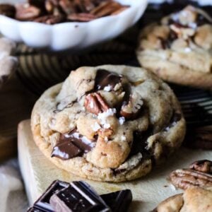 close up view of a thick cookie studded with pecans and chocolate chunks and sprinkled with flaky sea salt on a wooden cutting board.